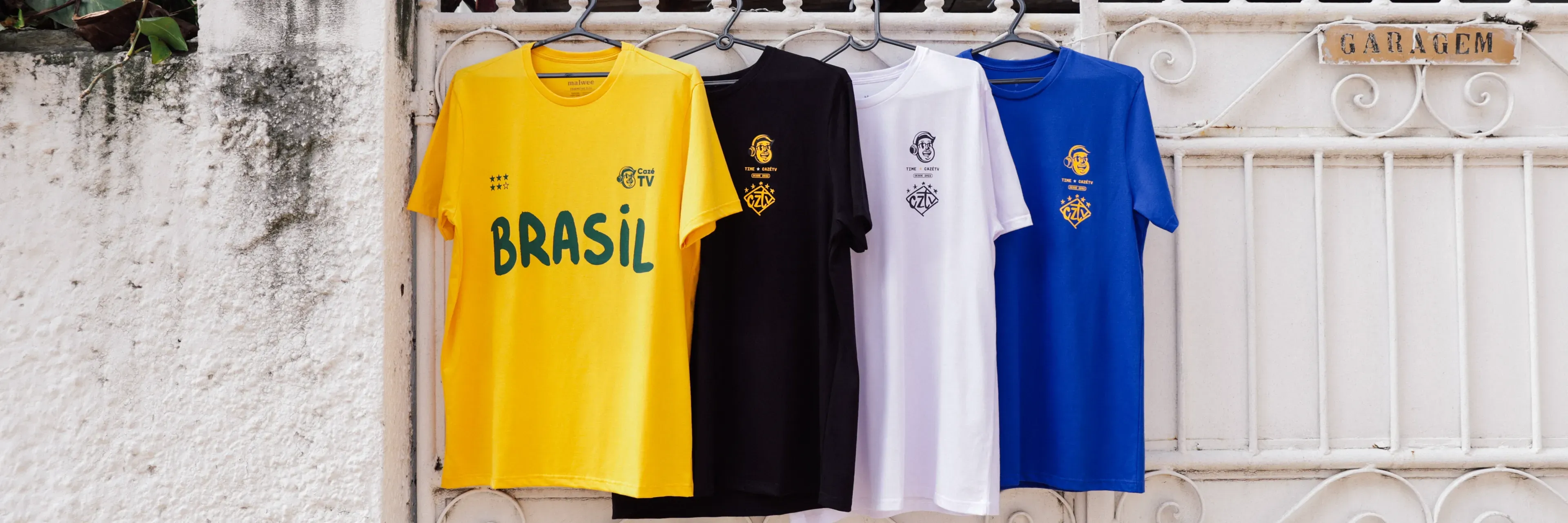 Coleção completa de camisas CazéTV Copa 2026 penduradas no varal: amarela, preta, branca e azul